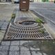 Sidewalk along Independence Street between 8th and 9th Streets in Shamokin, PA, showing deteriorated brick pavers with cracks, uneven surfaces, and debris. In the foreground, a partially broken metal tree grate surrounds a small pit with weeds growing through openings. A rusted trash can is near the curb. The street and intersection at 8th Street appear ahead.
