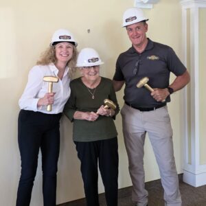 Three individuals wearing construction hard hats and holding ceremonial golden hammers during a preservation project kickoff event inside a historic building.